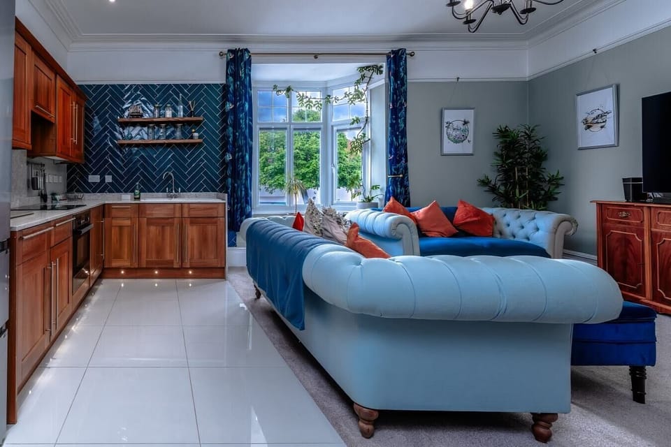 - Living room with blue sofas kitchen area and stylish wall design   - Bay window filling the space with daylight and greenery view   - Modern setup with Netflix TV perfect for meals and relaxation  