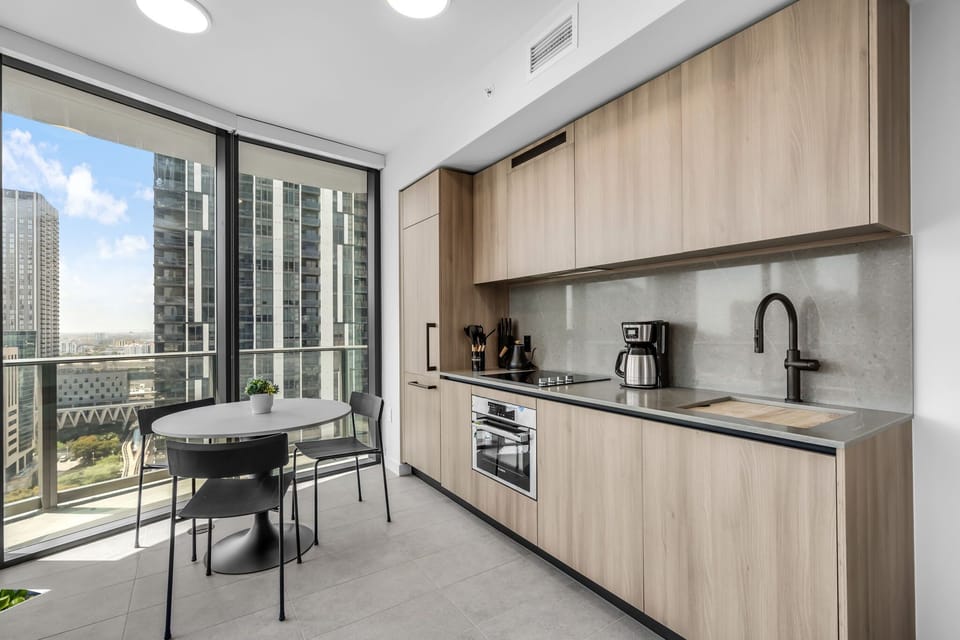 Cook and dine with a view! This sleek modern kitchen features light wood cabinetry and opens to a sun-drenched dining area with floor-to-ceiling windows. A perfect spot for home-cooked meals with a stunning city backdrop.