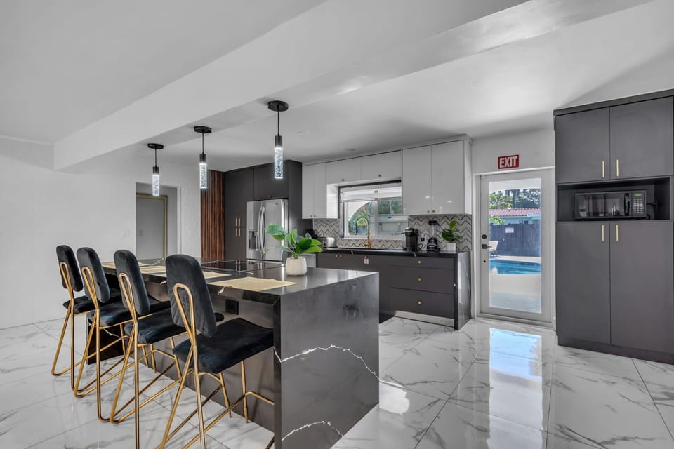 A chef's dream! Our bold luxury kitchen boasts a dramatic black marble island, velvet gold bar stools, crystal pendants, stainless appliances & a herringbone backsplash. Pool views too! 