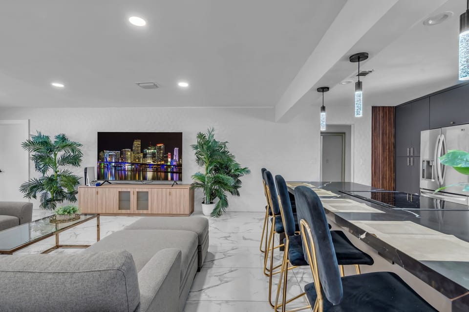 Where luxury meets comfort — black marble island with velvet gold bar seating flows into a bright living room with giant smart TV, lush palms & gleaming marble floors. Simply stunning! 