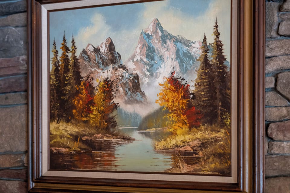 A framed landscape painting depicting snow-capped mountains, autumn foliage, and a tranquil stream—adding a cozy, artistic touch to the space.