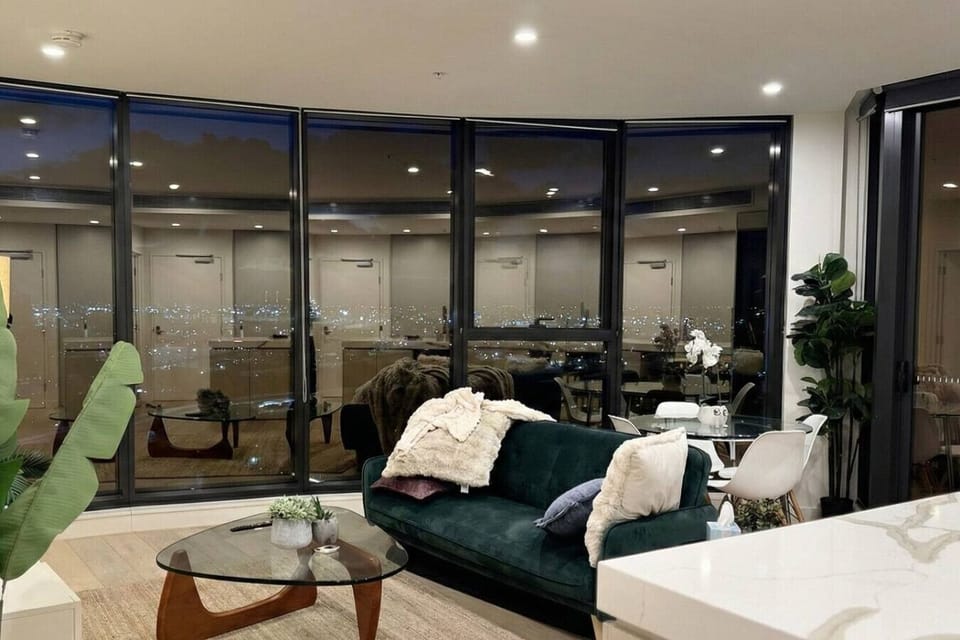 Curved living room at night with a deep green velvet sofa, glass coffee table, and indoor plants, wrapped by floor-to-ceiling windows reflecting the glittering city lights spread across the horizon.