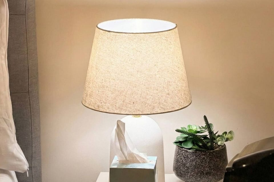 A bedside table with a warm linen-shade lamp and small potted succulent, casting a soft glow beside the bed. A simple, calming setup that makes winding down at the end of the day easy and comfortable.