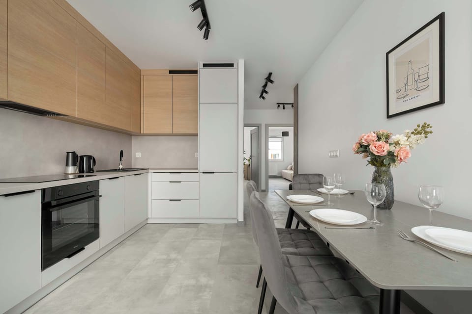 The kitchen perspective shows a long countertop extending toward a dining table in the distance. White tiled floors and bright walls enhance the sense of space.
