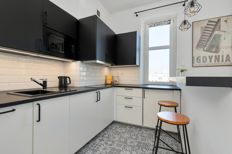 A bright kitchenette area shows white lower cabinets paired with black upper units and a white subway tile backsplash. Two wooden stools are placed next to a breakfast bar, and patterned floor tiles add a vintage touch.