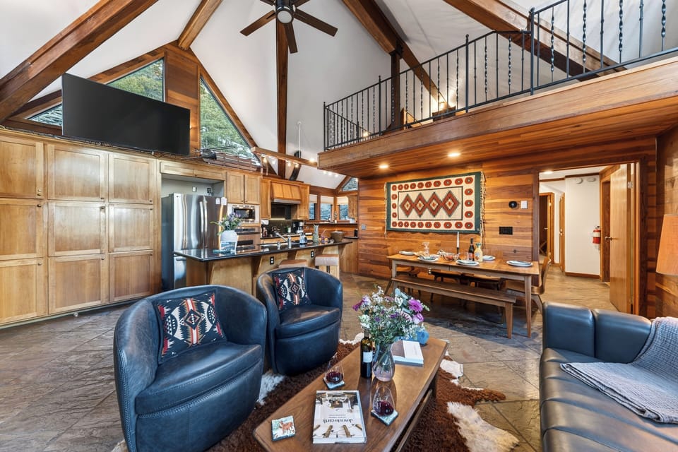 Cozy cabin living and dining space with warm wood tones, vaulted windows, and a stone fireplace anchoring the room.
