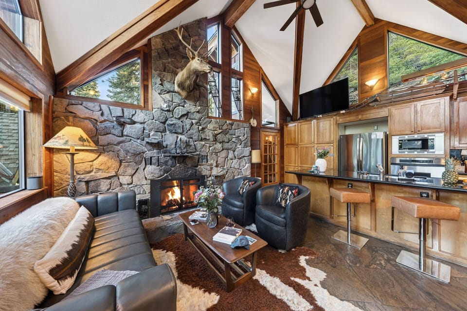Cozy cabin living and dining space with warm wood tones, vaulted windows, and a stone fireplace anchoring the room.