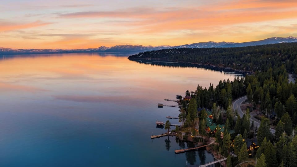 “Sunset over a glass‑calm lake framed by evergreens and distant snow‑capped peaks.