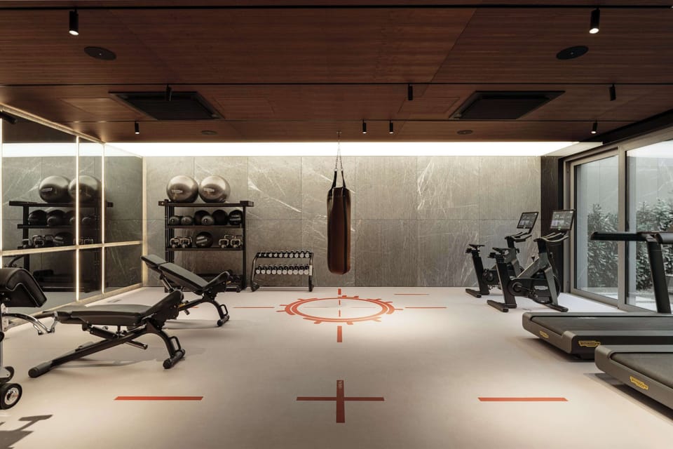 A fully equipped fitness center featuring weight benches, cardio equipment, and a heavy boxing bag hanging in the center. Red cross markings on the floor define individual training zones, while large glass walls bring in ample light.