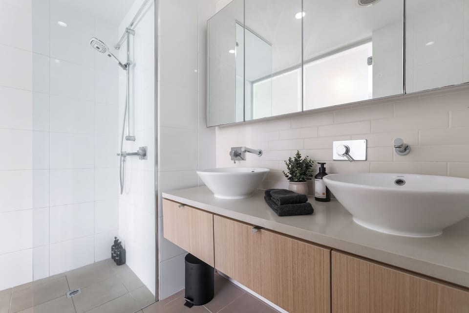A bright bathroom with twin vessel sinks on a timber vanity and a mirrored cabinet above for storage. A glass-enclosed shower with a handheld head makes getting ready easy, even when two guests share the space.