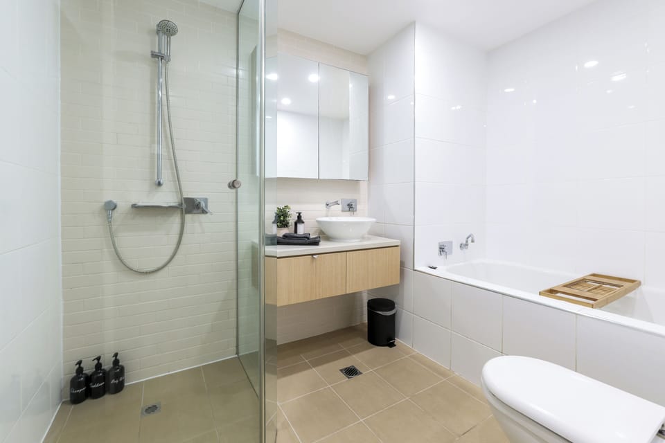 A clean, bright bathroom with a glass-enclosed shower and handheld head, a timber vanity with vessel sink, and a mirrored cabinet for storage. A full bathtub gives guests the choice of a quick rinse or a longer soak.