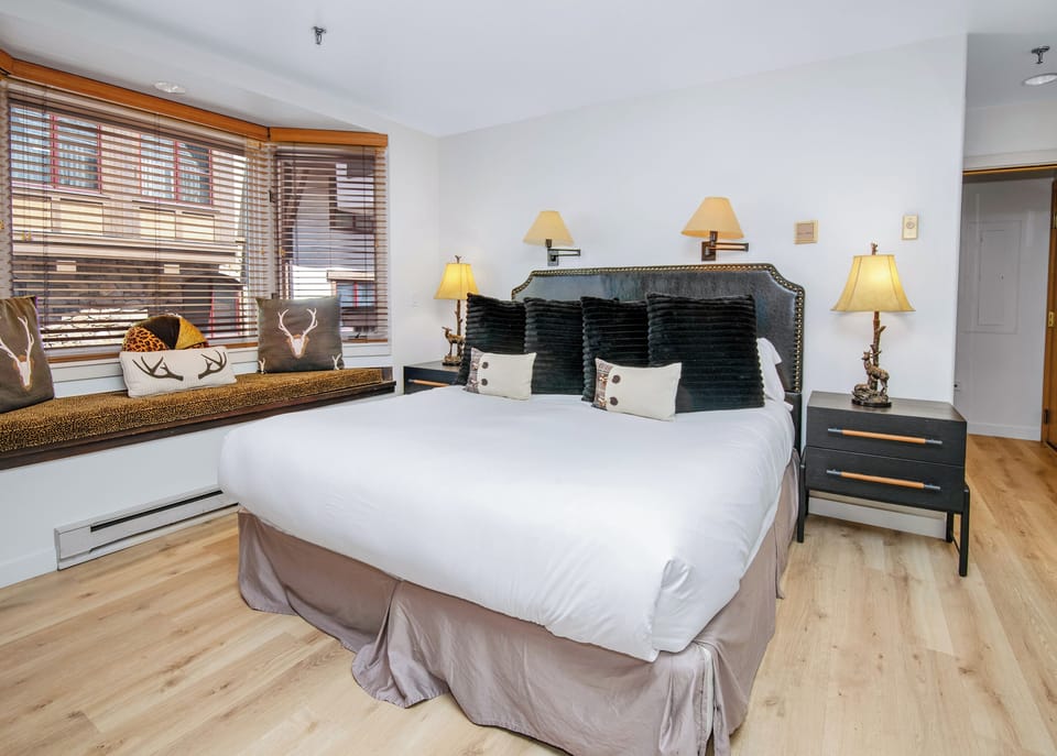 Your cozy bedroom features a plush bed with stylish linens, warm lighting, and charming alpine touches. Wooden blinds filter soft natural light, while elegant nightstands complete the inviting space.