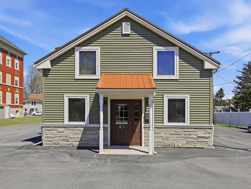 The straight-on daytime exterior shows the green two-story building, covered central entrance, metal porch roof, four front windows, gray stone-look lower facade, and paved frontage.