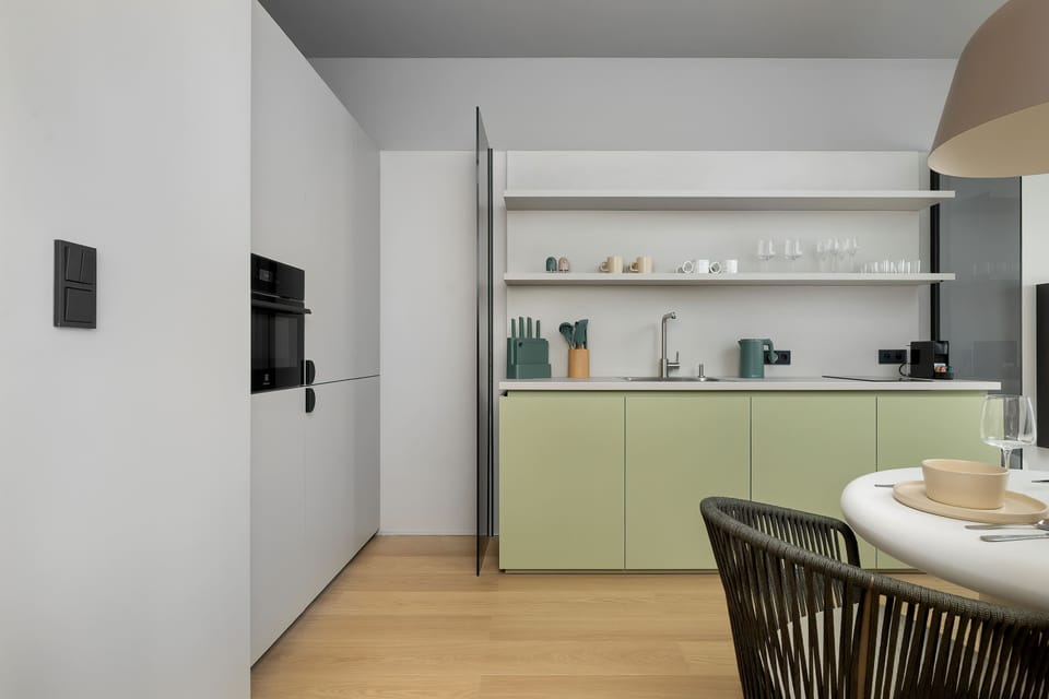 A view of a minimalist kitchen with light sage green and white cabinets, featuring integrated appliances, a small white dining table with an interesting textured edge, and woven-back chairs, set against light wood flooring and grey-blue walls.
