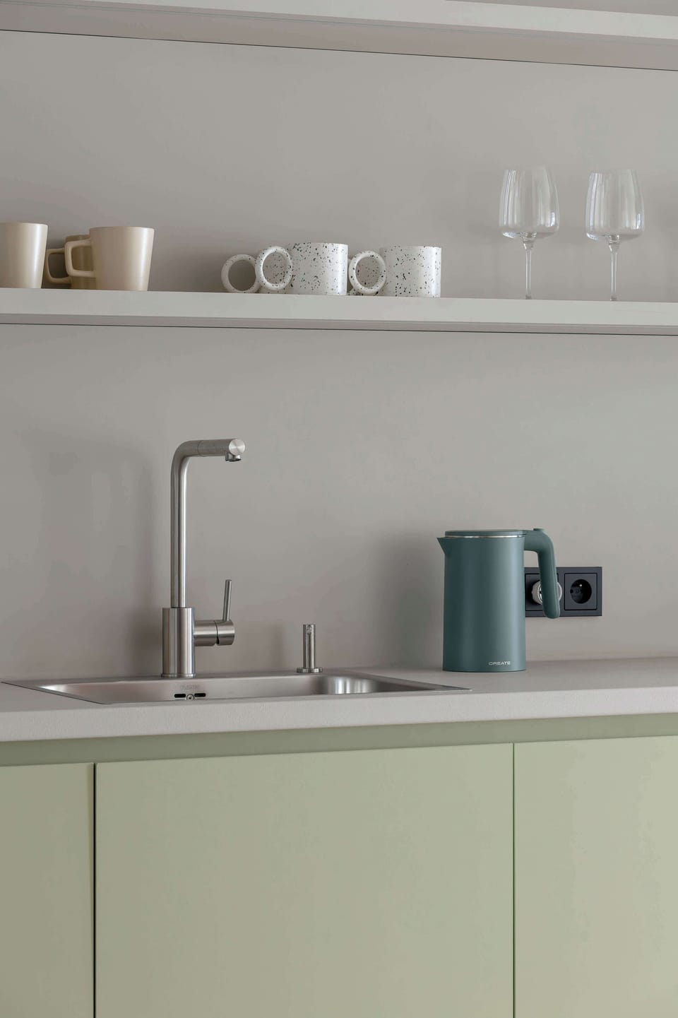 A focused close-up on the kitchen countertop area, highlighting a modern stainless steel faucet and sink, with a green kettle and several white mugs and a glass arranged neatly on the shelf above.