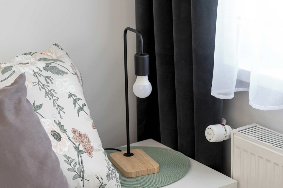 A minimalist bedside setup includes a wooden lamp base with an exposed designer bulb. The floral pattern of the pillow and the white window curtain are visible in the background.