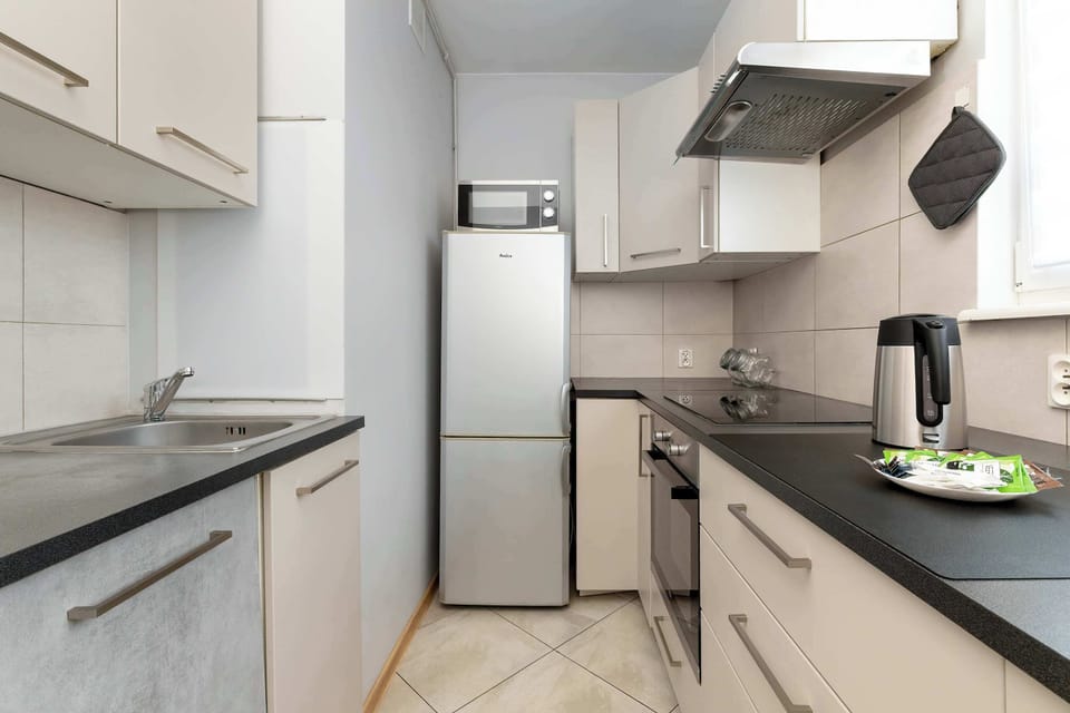 The compact kitchen is equipped with a tall silver refrigerator and white cabinetry. A black electric kettle sits on the counter next to a built-in stovetop and oven.