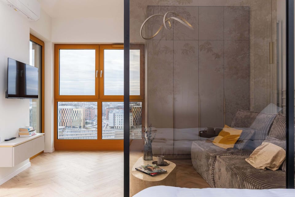 A view of the living room through the bedroom’s glass partition, which emphasizes the apartment’s open character. Warm afternoon sunlight streams in through the large window in the background.
