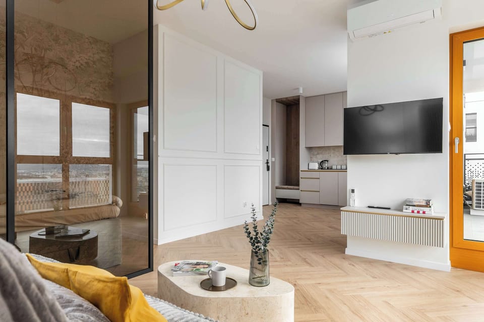 A view from the living room toward the kitchenette, showcasing the light, herringbone-patterned wooden floor. A TV is mounted on the wall, and next to it is an exit to the sunny terrace.