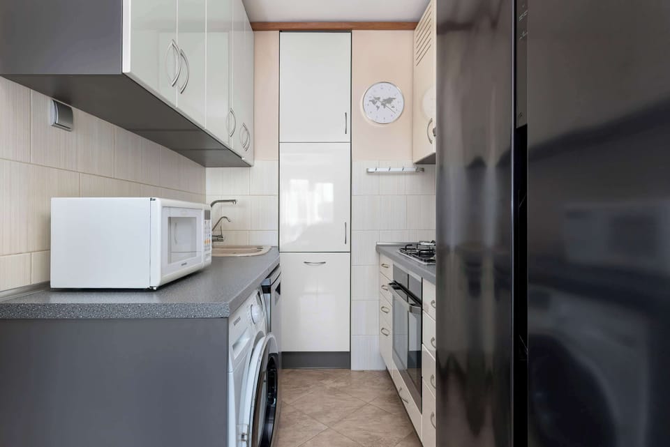 This perspective of the kitchen shows the proximity of the microwave to the tall white pantry cupboards. The dark refrigerator on the right contrasts with the bright white walls and cabinetry.