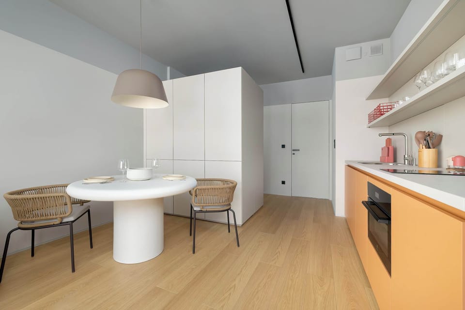 A bright, modern dining area with a round white table and minimalist chairs placed near a large window. The space connects seamlessly with a sleek kitchenette featuring light cabinetry and warm wooden accents. 