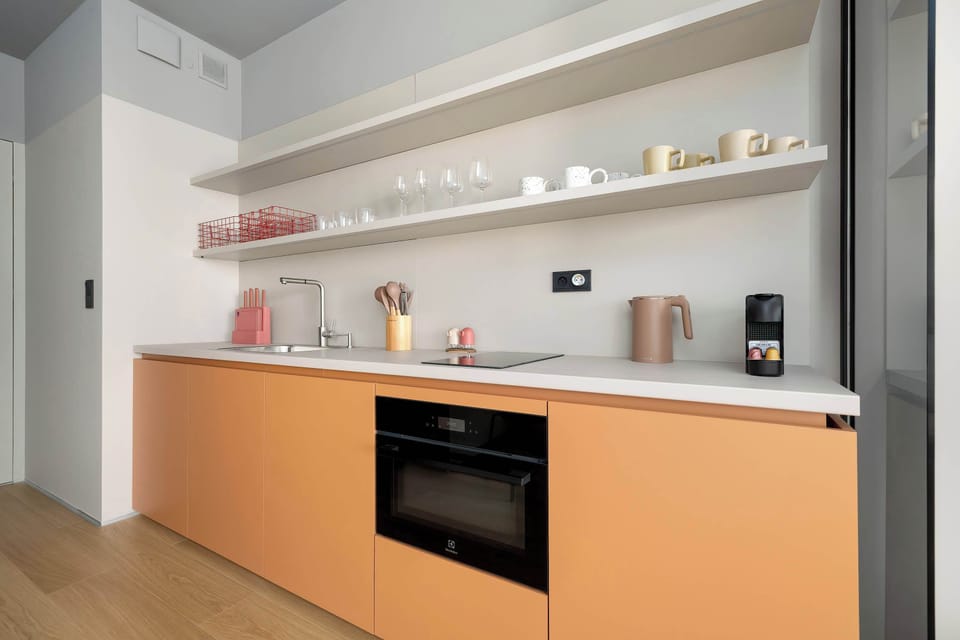 A compact kitchenette with a built-in oven, open shelving, and smooth white surfaces complemented by a wooden countertop. The design is simple and functional, with a few decorative items placed on the shelves.