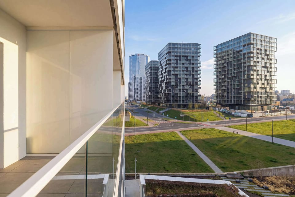 A balcony view overlooking a modern residential complex with green spaces and pathways. Large windows or glass doors frame the view, bringing in plenty of natural light. The surroundings appear peaceful and thoughtfully designed.