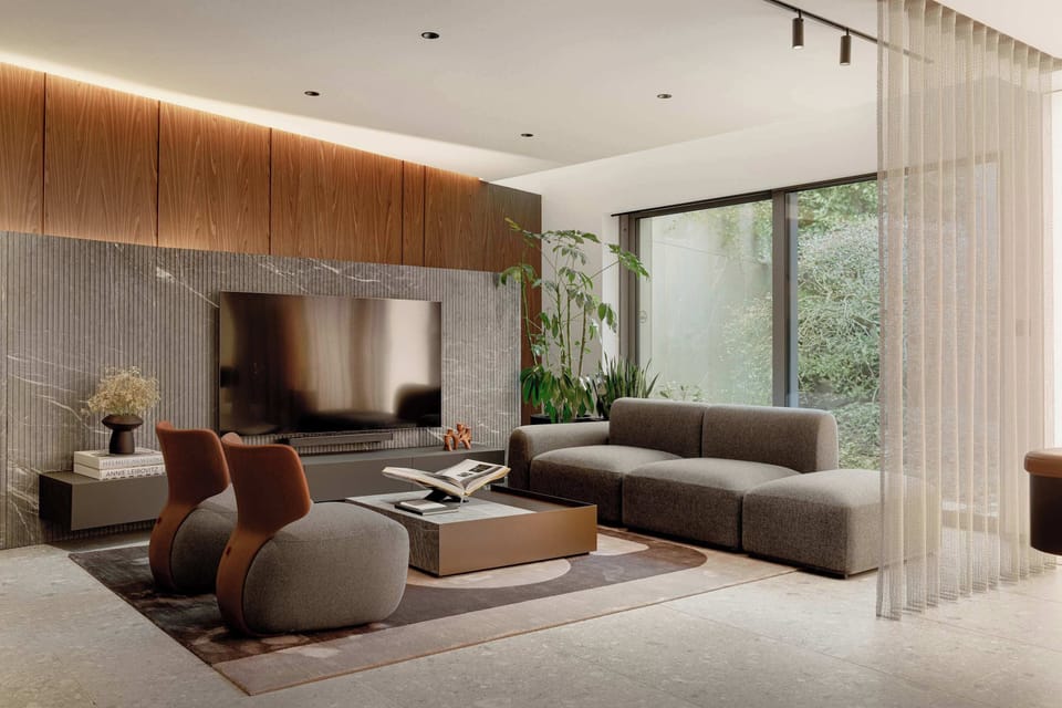A contemporary living room arranged with a plush sectional sofa and two accent armchairs facing a large mounted television. Warm wooden paneling accents the main wall, complemented by natural light from adjacent glass doors.