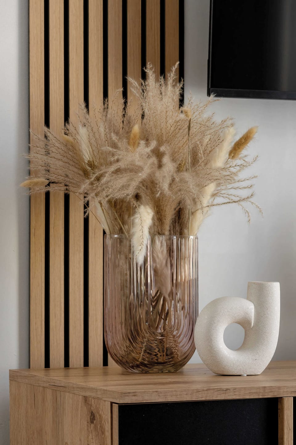 A detail shot showing a light wood media console with stylized vertical slatted panels. A glass vase holds a large arrangement of dried pampas grass and other botanicals.