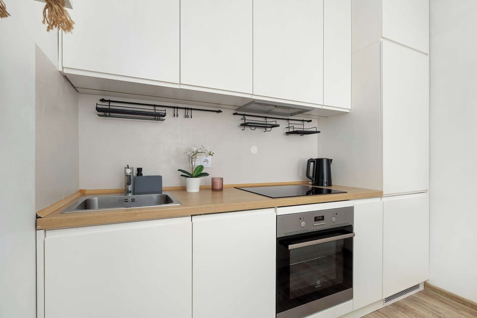 A modern kitchen with minimalist white cabinetry and light-colored countertops. A built-in oven and a coffee machine are visible on the counter.