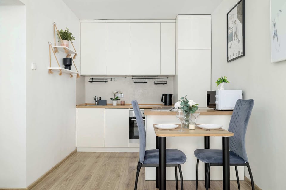 A compact, open-plan kitchen and dining area featuring clean white cabinets, light wood countertops, and two blue upholstered dining chairs. The room has a clear view into a living space beyond.