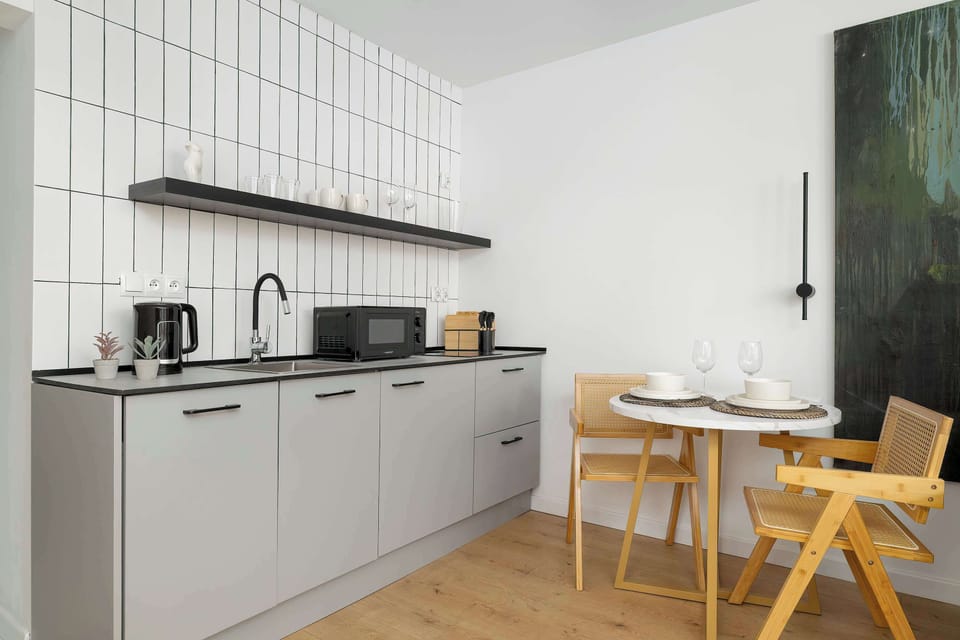 The kitchenette area is designed with light grey cabinets and a black countertop, complemented by a white tiled backsplash. A small wooden dining table with two chairs is tucked neatly into the corner.