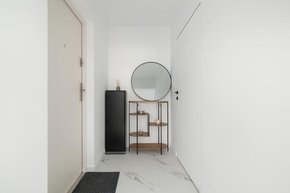 The entryway is bright and minimalist, featuring a white door and a large round mirror above a wooden console table. Marble-effect floor tiles create a luxurious first impression.