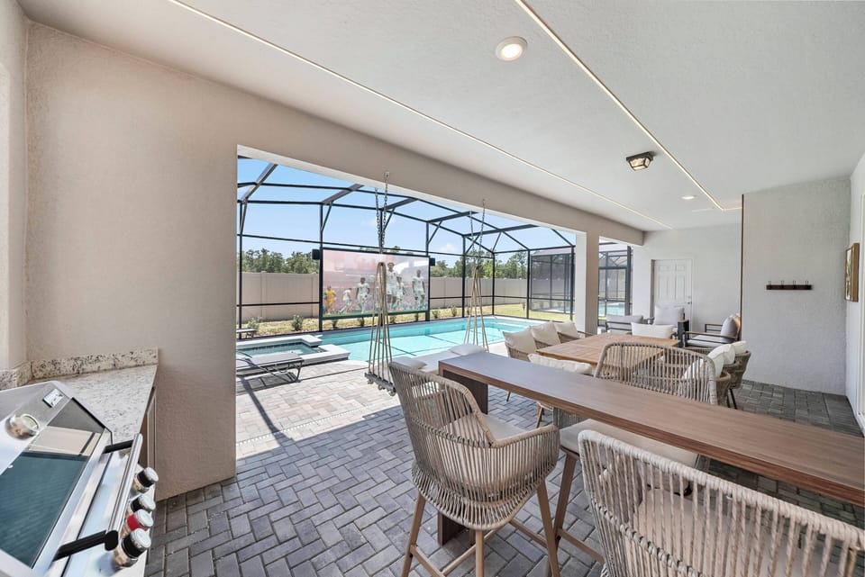This airy dining area features a sleek wooden table surrounded by comfortable woven chairs, perfect for meals with a view. Large screens frame the pool outside, inviting you to enjoy sunny days and relaxing evenings in style.