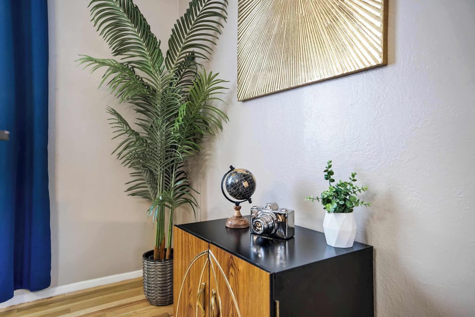 A tall indoor palm beside the media console brings life and texture to the corner, complemented by a geometric gold wall piece and a small collection of curated objects on the console. The navy window panel behind adds a rich contrast to the warm woo
