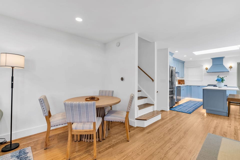 Dining nook with round wood table and striped upholstered chairs for four, plus open view of the staircase and blue-cabinet kitchen beyond.