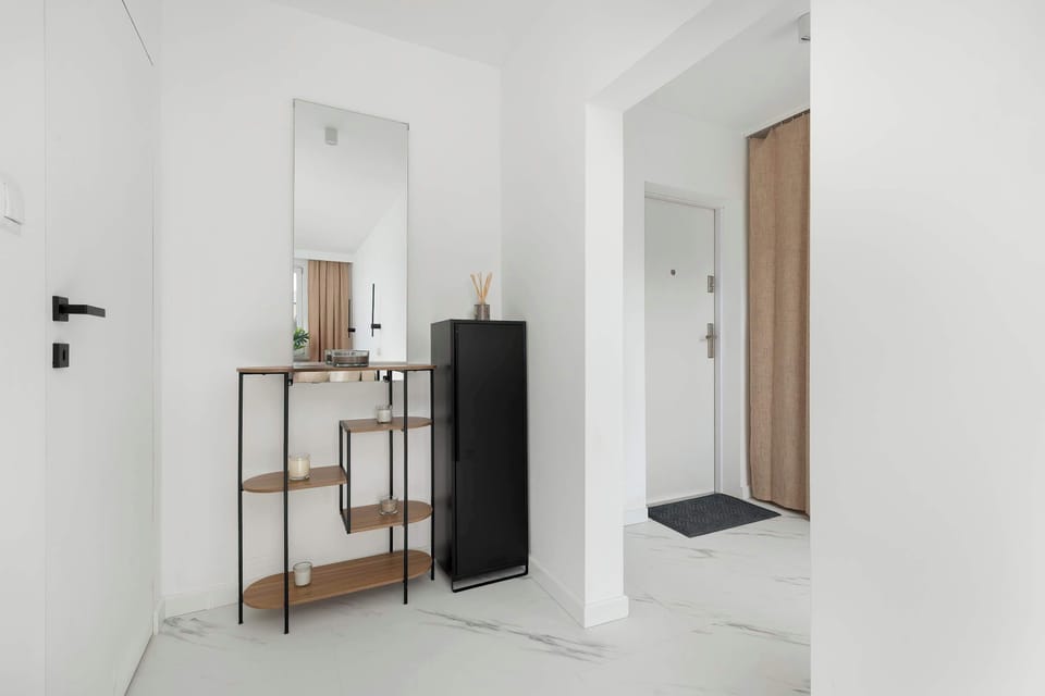 A bright hallway with a minimalist design and light flooring. A simple shelving unit provides storage and decorative space, while the overall look remains uncluttered. The area feels open and welcoming.