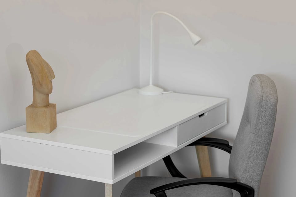 A dedicated home office nook features a clean white desk, an ergonomic chair, and a bright desk lamp. A unique wooden sculpture adds an artistic touch to this quiet corner of the apartment.