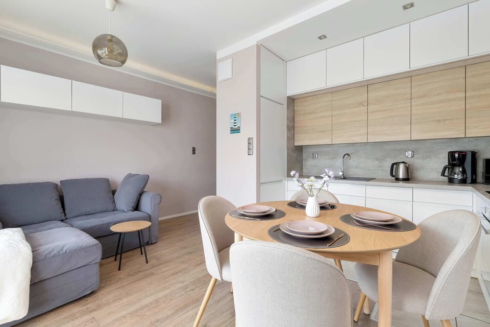 Looking from the living room toward the kitchen, the layout reveals a spacious and social floor plan. The neutral palette of the grey sofa and white walls is complemented by light wood furniture.