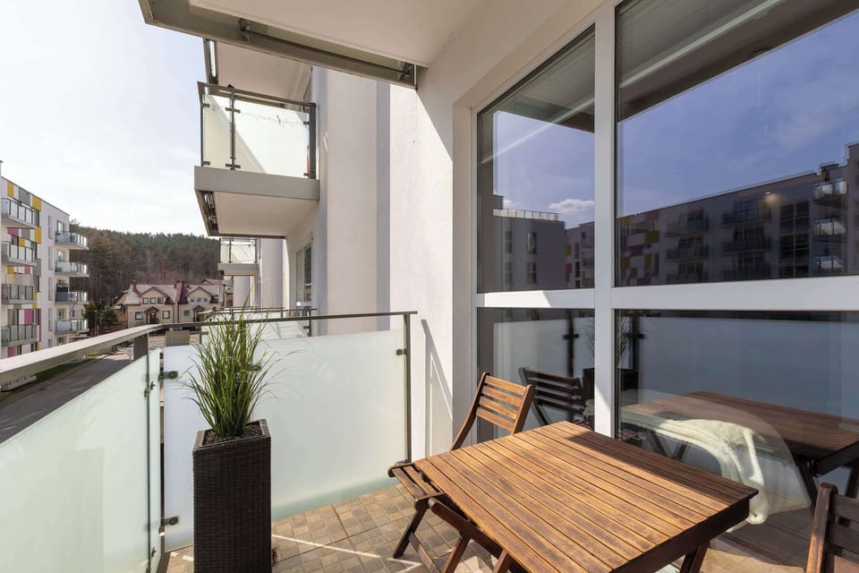The private balcony is furnished with a wooden table and chair, offering a perfect spot for morning coffee. A glass railing ensures an unobstructed view of the nearby water and surrounding architecture.