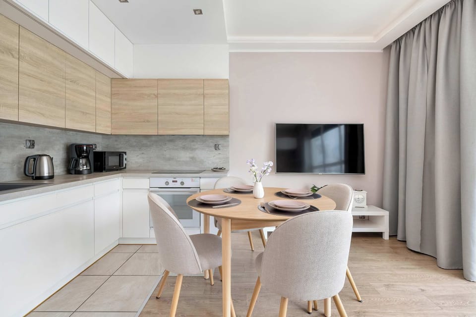 The perspective from the kitchen area highlights a bright dining corner with four white chairs around a wooden table. Large windows in the background provide a view of the outdoors and keep the interior airy.