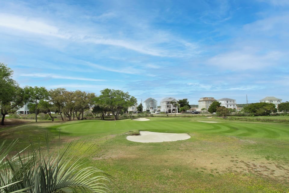 A pristine golf course with manicured greens and sand bunkers, surrounded by coastal homes and natural vegetation.