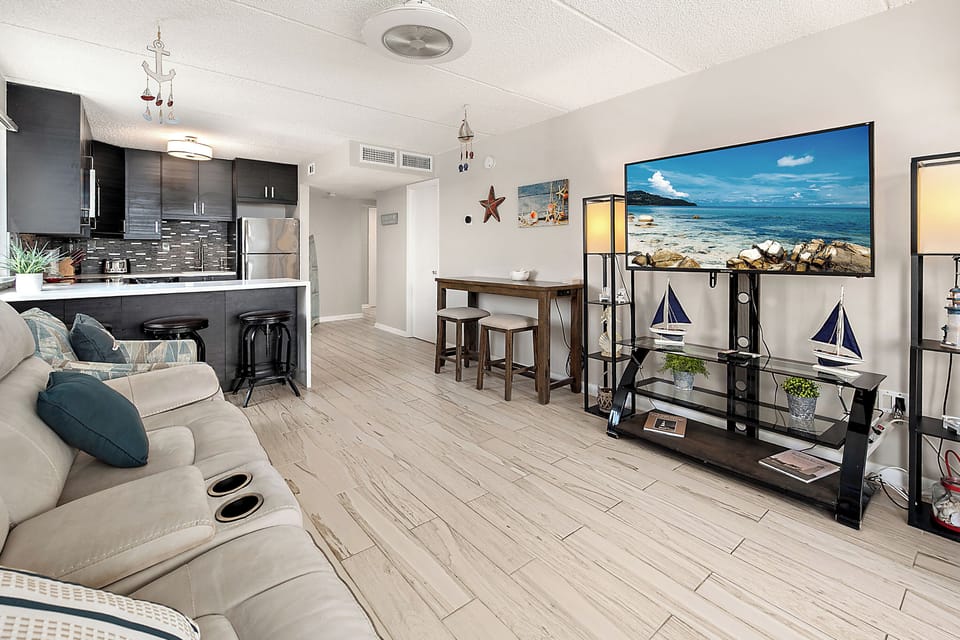 One room, everything you need. A spacious open-concept living space with plush seating, a large TV, coastal decor, and a fully equipped kitchen just steps away