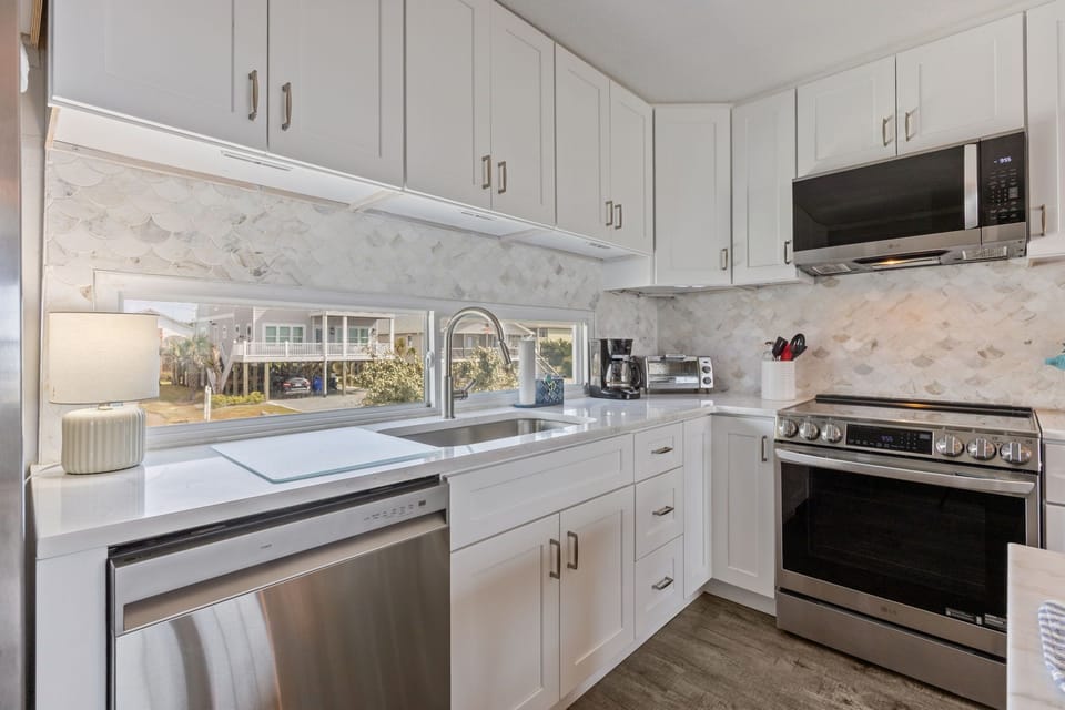 Bright white kitchen with stainless appliances, marble-look table, six chairs, modern lighting, wood-look floors, and stair access nearby.
