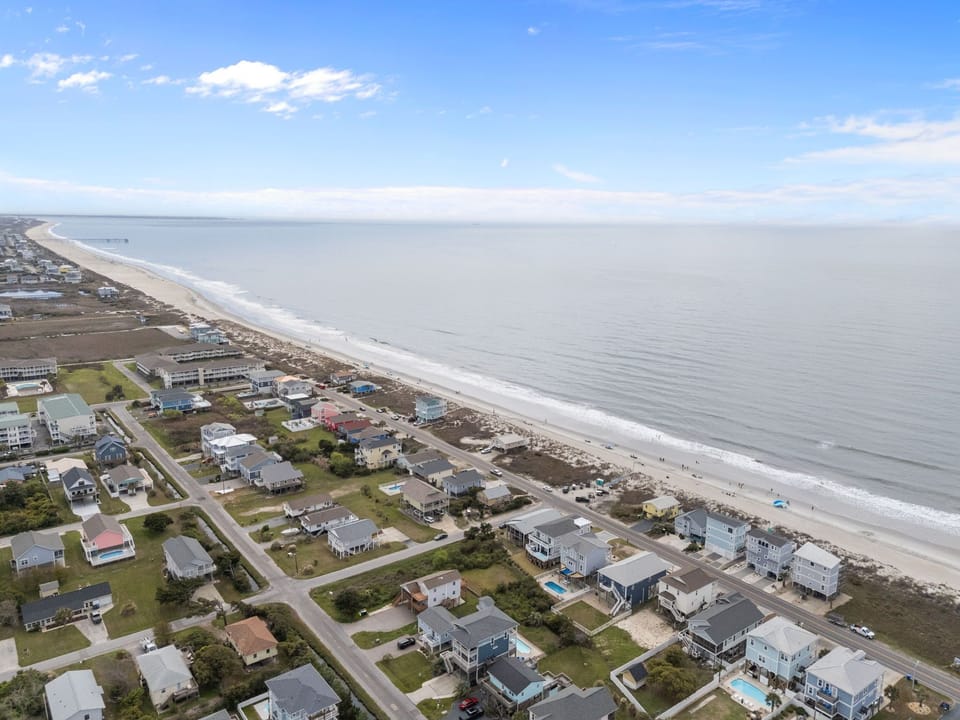 Aerial view of a marked oceanfront home on Oak Island with neighboring beach houses, road frontage, sandy dunes, wide shoreline, and expansive Atlantic Ocean views.