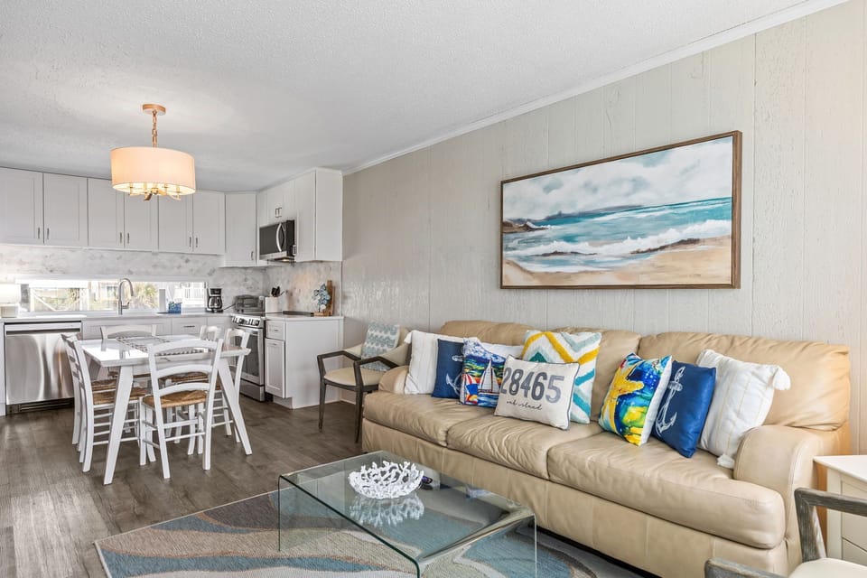 Coastal living room with a fireplace, wall-mounted TV, glass coffee table, accent chair, beachside windows, door to the deck, and soft ocean-view cottage style.