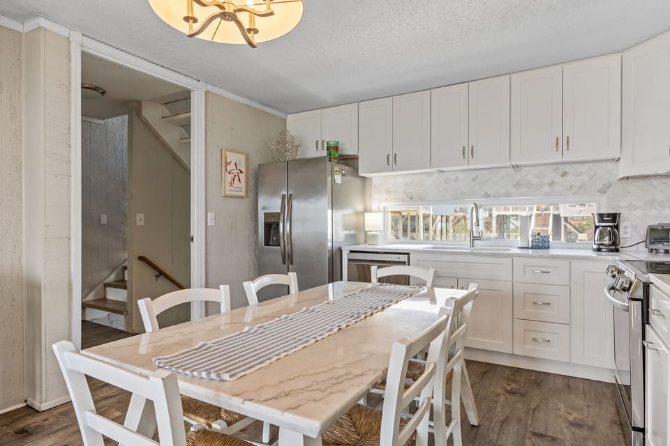 Bright white kitchen with stainless appliances, marble-look table, six chairs, modern lighting, wood-look floors, and stair access nearby.