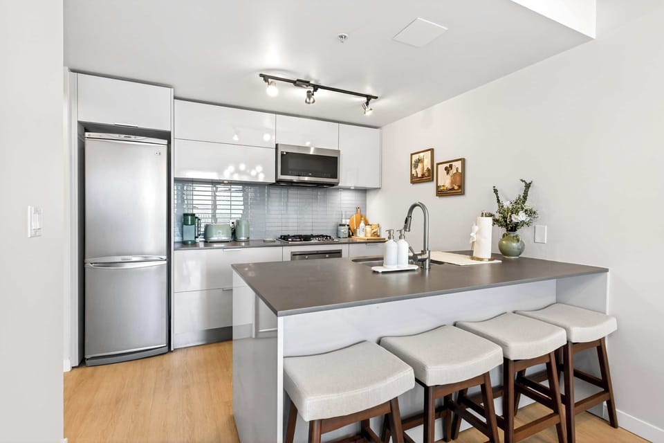 Contemporary apartment kitchen design with high-gloss white cabinetry, stainless steel appliances, grey countertops, and a functional breakfast bar peninsula with wooden stools.