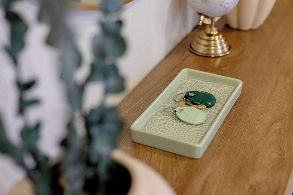 A sage green ceramic key tray with an intricate floral pattern, holding two leather key fobs in dark forest green and pale mint on a wooden console table.