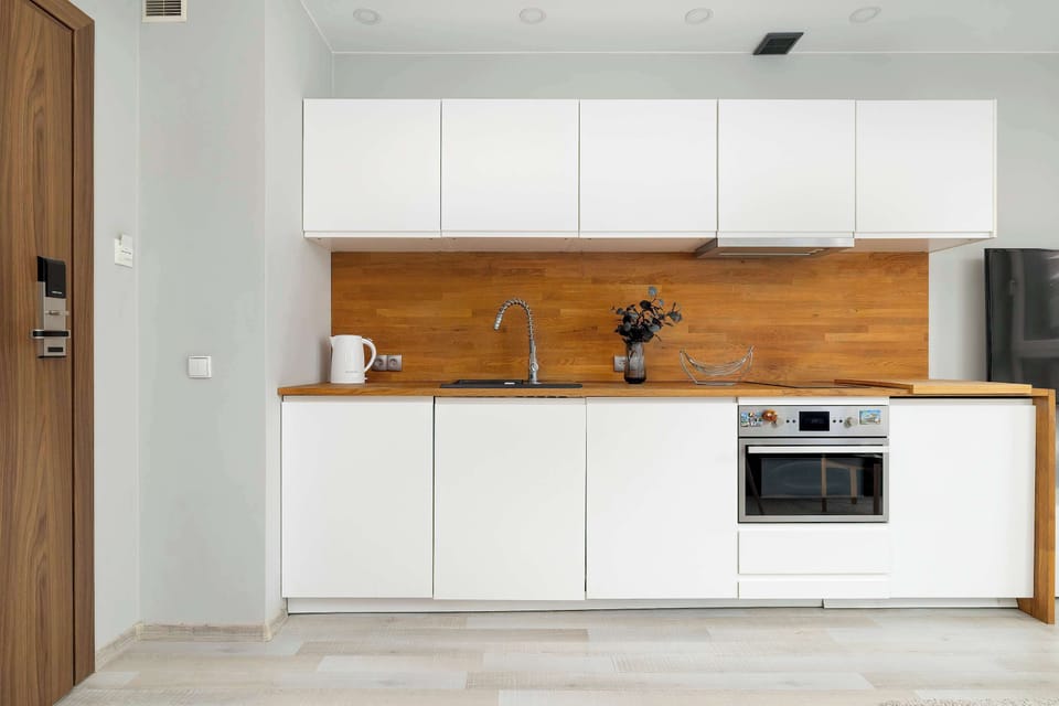 A sleek, linear kitchenette showcases white cabinetry contrasted by a warm, natural wood backsplash and countertop. The space is equipped with modern integrated appliances including a stove and oven.